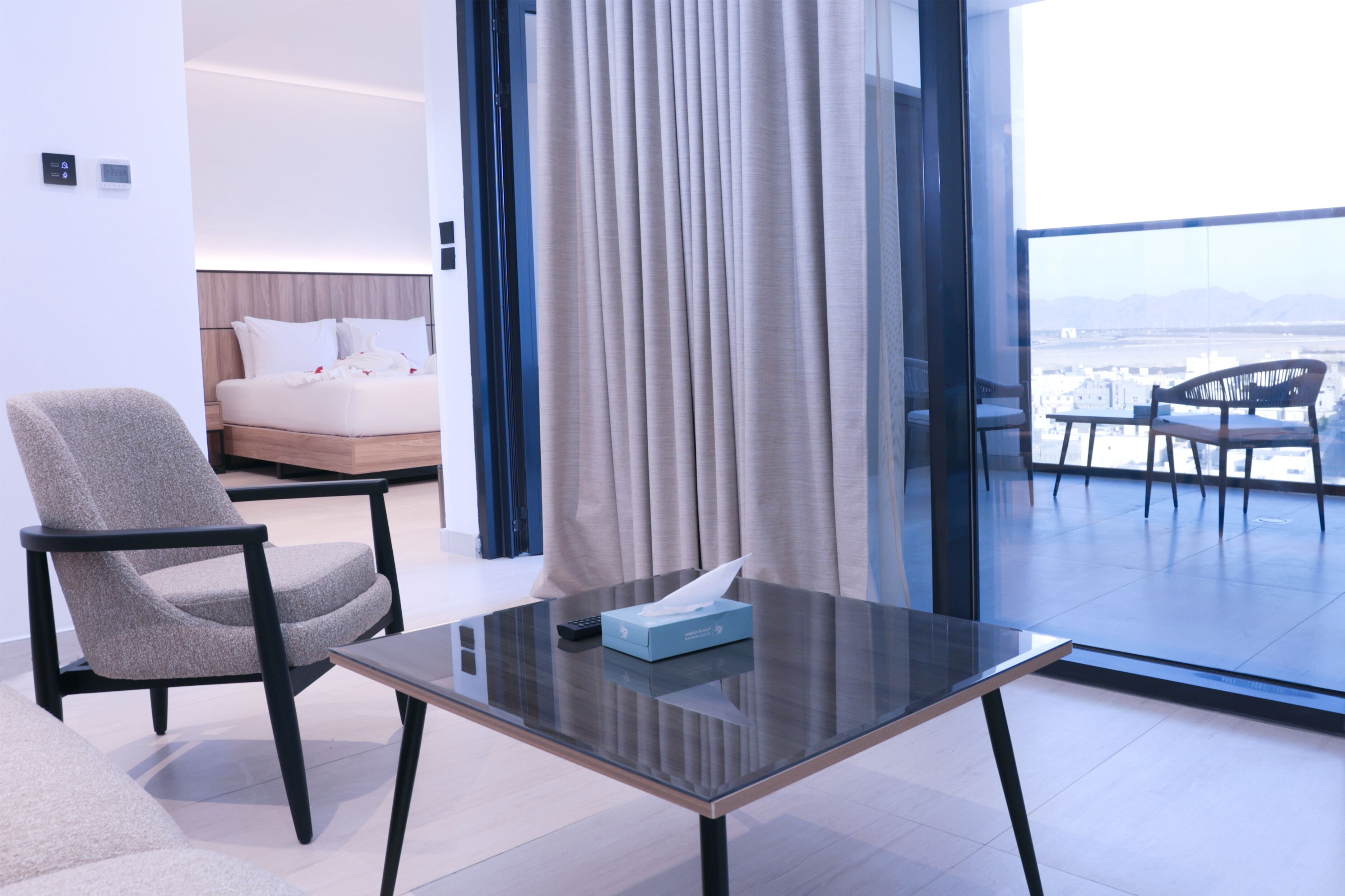 Una camera d'albergo moderna con un tavolino in vetro, un divano, un letto con lenzuola bianche e vista sulla città attraverso ampie finestre.