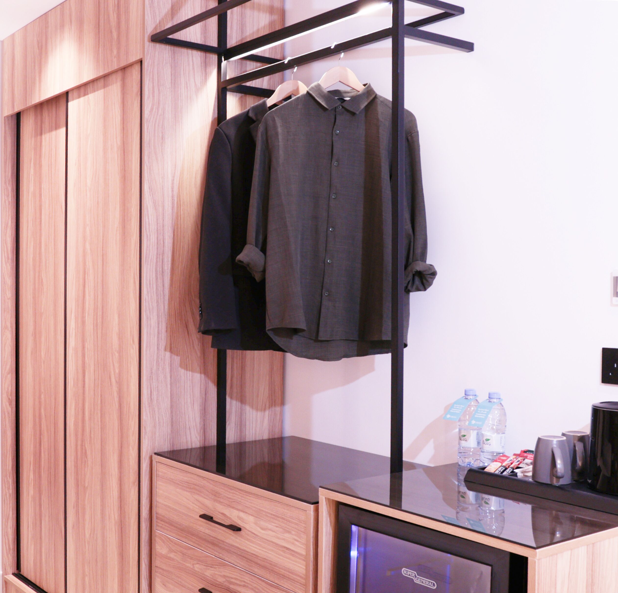 A modern hotel room with a wooden wardrobe, a black desk with a coffee machine, and a hanging rack with two dark shirts.