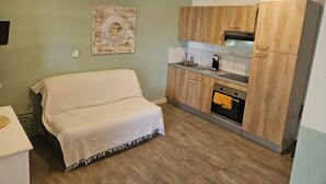 Une cuisine de taille réduite, avec des meubles en bois, un évier et une cuisinière. La pièce adjacente contient un lit recouvert d’une couverture blanche.