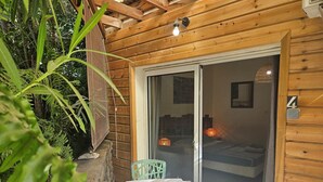Una cabaña de madera con un cerramiento de vidrio corredizo que da a un dormitorio. En el exterior, hay una mesa con una bandeja de madera y dos sillas verdes.