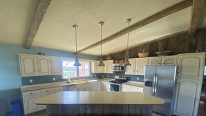 Una cocina con una isla grande, taburetes de madera, armarios blancos, un refrigerador de acero inoxidable y una lámpara colgante.