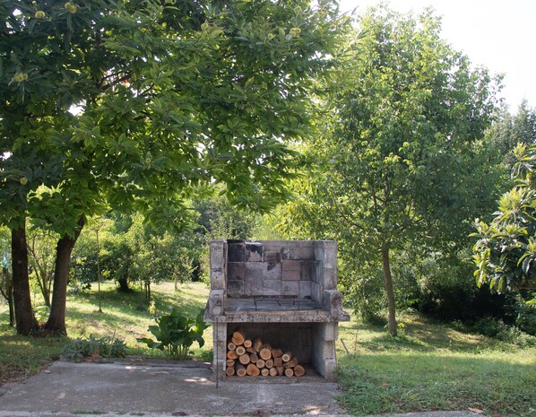 Una chimenea de concreto con una pila de leña en su interior, rodeada de árboles y césped verdes y frondosos.