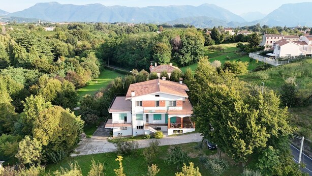 A two-story house with a tiled roof, surrounded by lush greenery and mountains in the background.