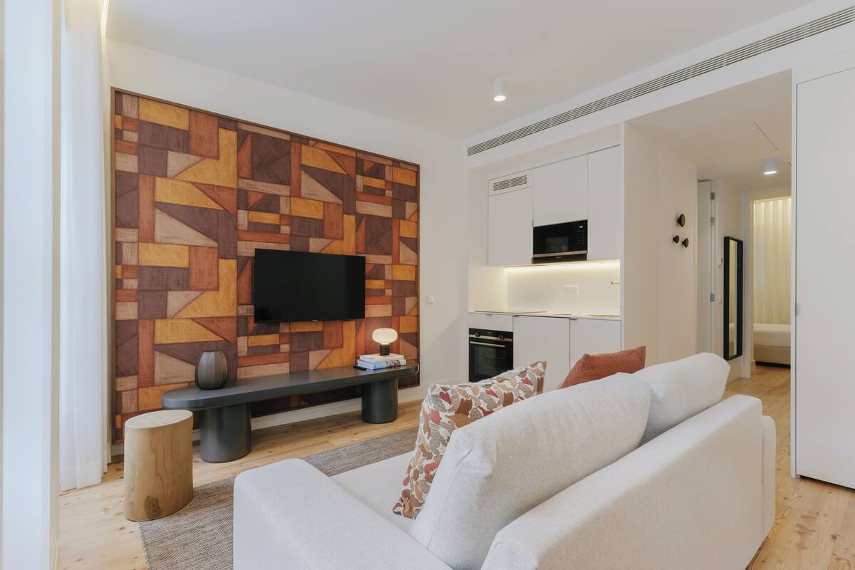 A modern living room with a flat-screen TV mounted on a geometric-patterned wall, a sofa with patterned cushions, a small round wooden stool, and a bench with a lamp.