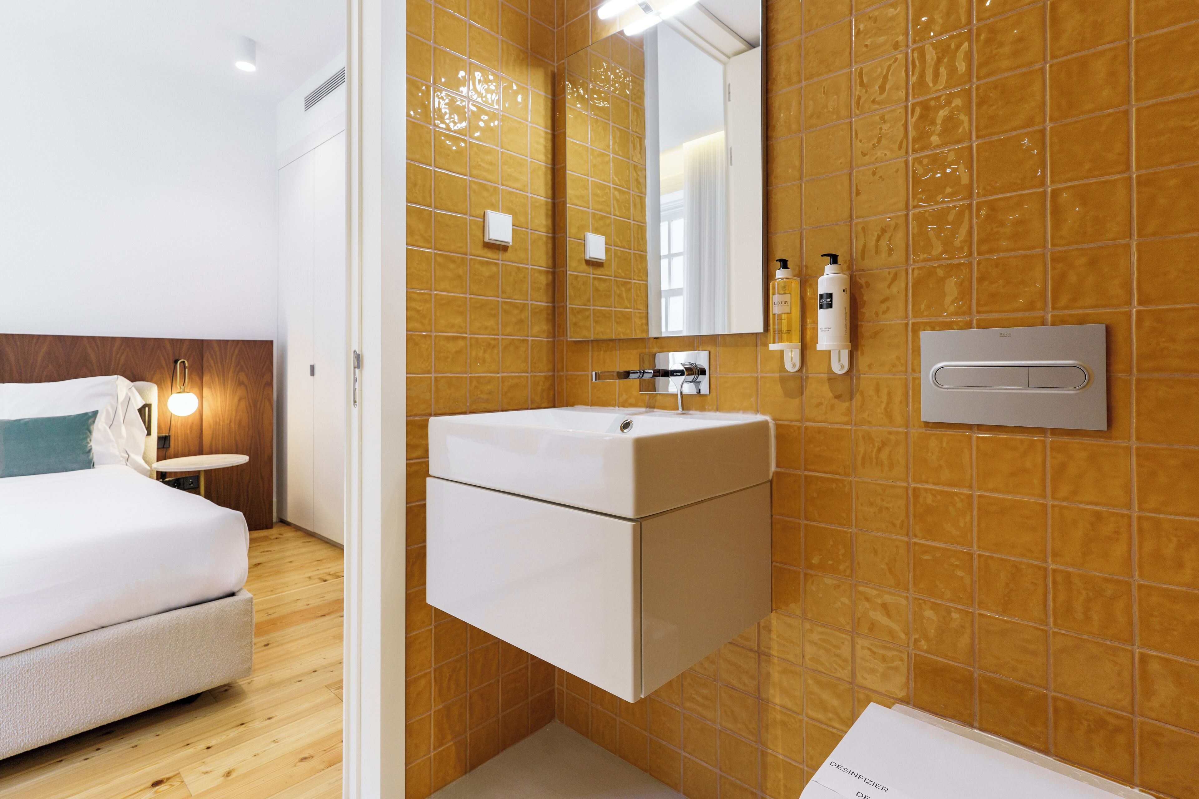 A modern hotel room with a wooden floor, a white bed, a built-in white sink, and a mirror above it.