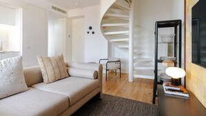 En moderne stue med sofa, lænestol, trappegang i spiral og et sofabord med bøger og en lampe.