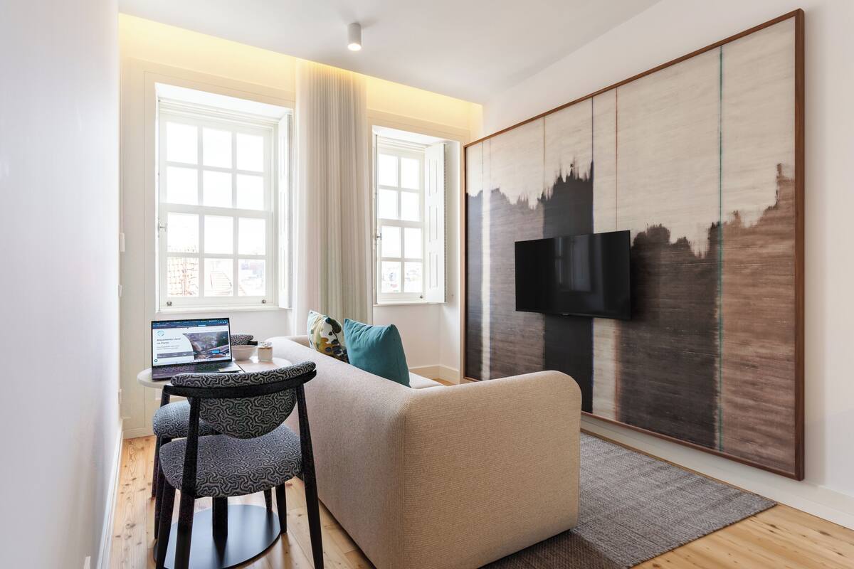 A modern living room with a sofa, a coffee table, and a laptop. There is a large artwork on the wall and a TV mounted above it.