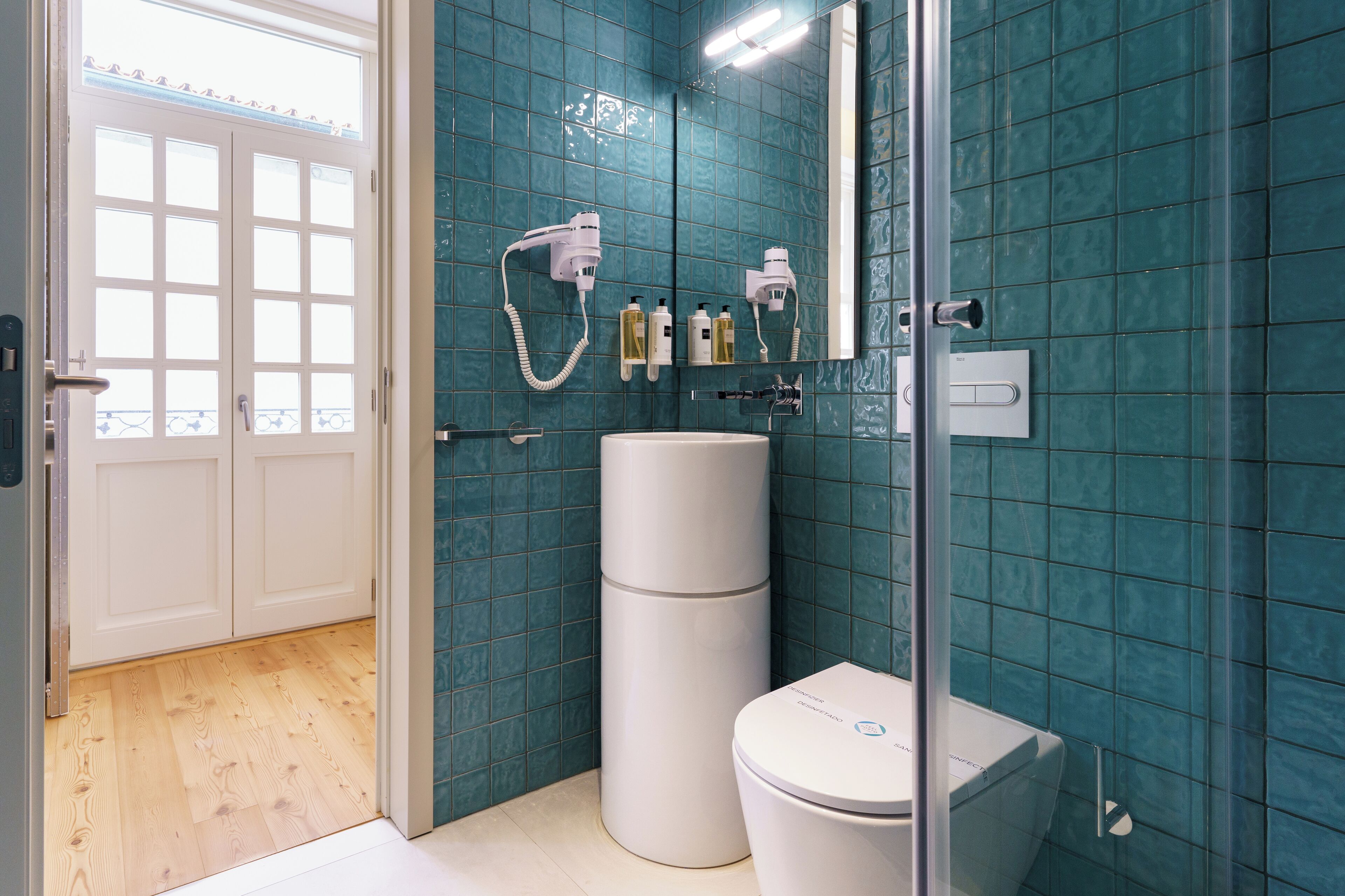 Een badkamer met blauwgroene tegels, een witte wastafel en een toilet. Er hangen twee haardroger aan de muur.