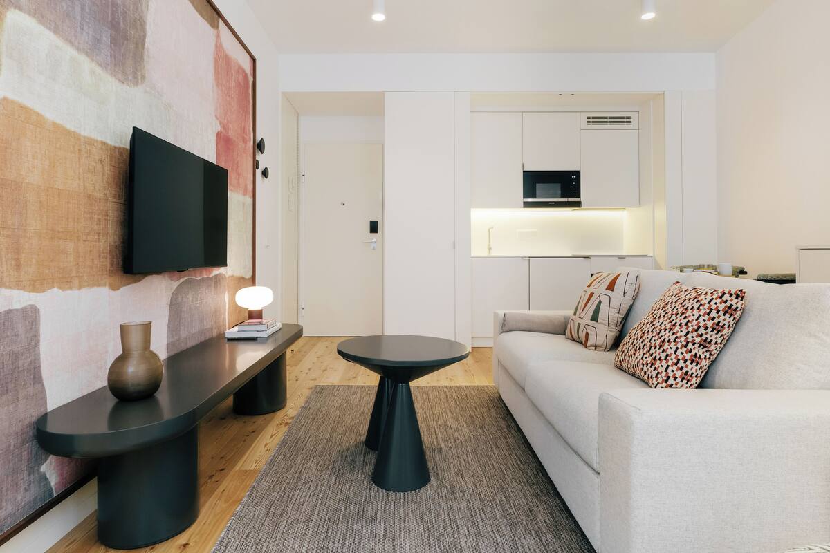A modern living room with a flat-screen TV mounted on a textured wall, a round black coffee table, a sofa with patterned cushions, and a kitchen area in the background.