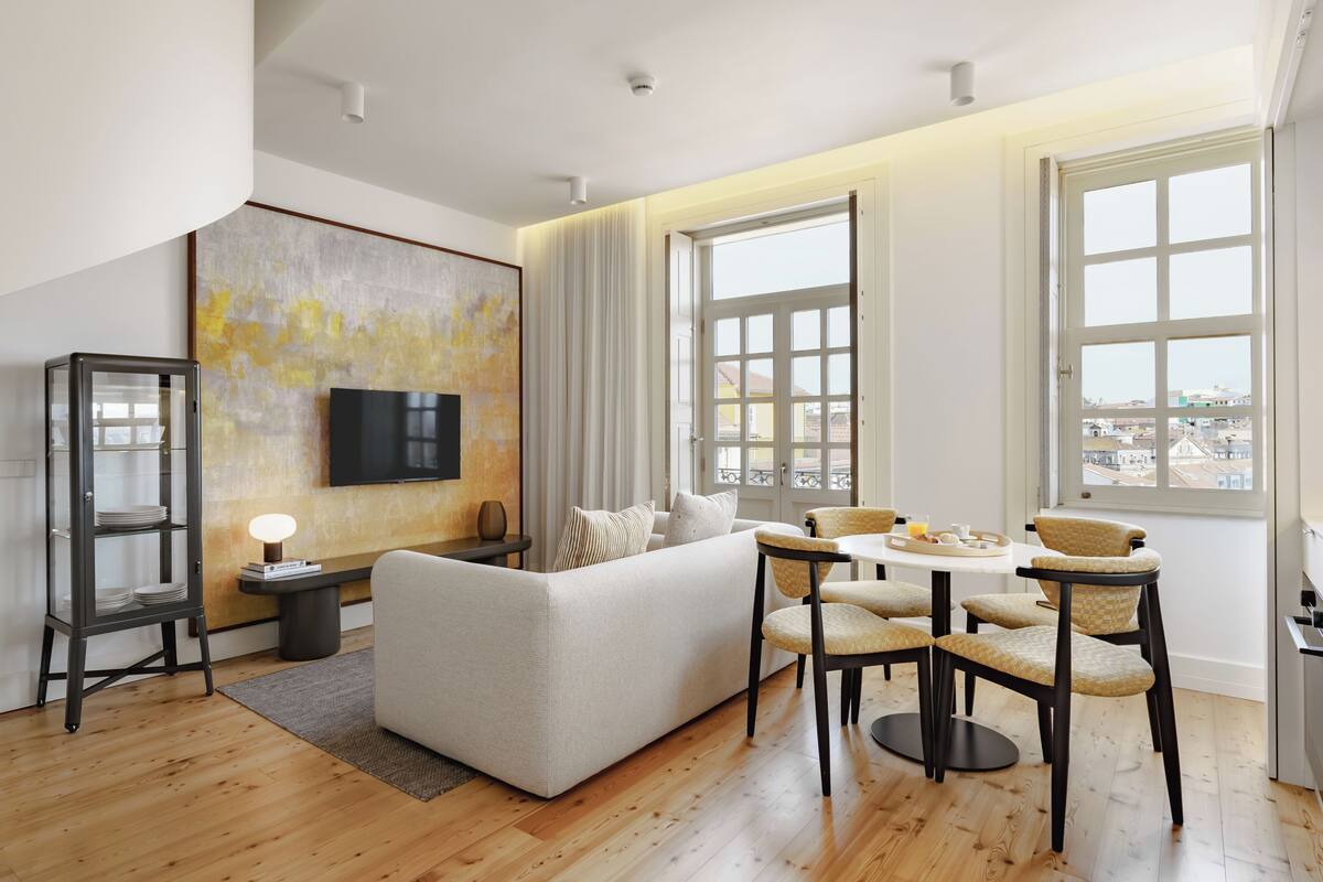 A modern living room with a sofa, dining table, and chairs, large windows with city views, and a flat-screen TV on the wall.