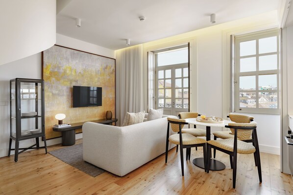 Un soggiorno moderno con divano, tavolo da pranzo e sedie, ampie finestre con vista sulla città e una TV a schermo piatto appesa al muro.