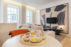 Una moderna camera d'hotel con angolo ristoro, colazione servita, divano, televisore a schermo piatto e un grande quadro astratto.