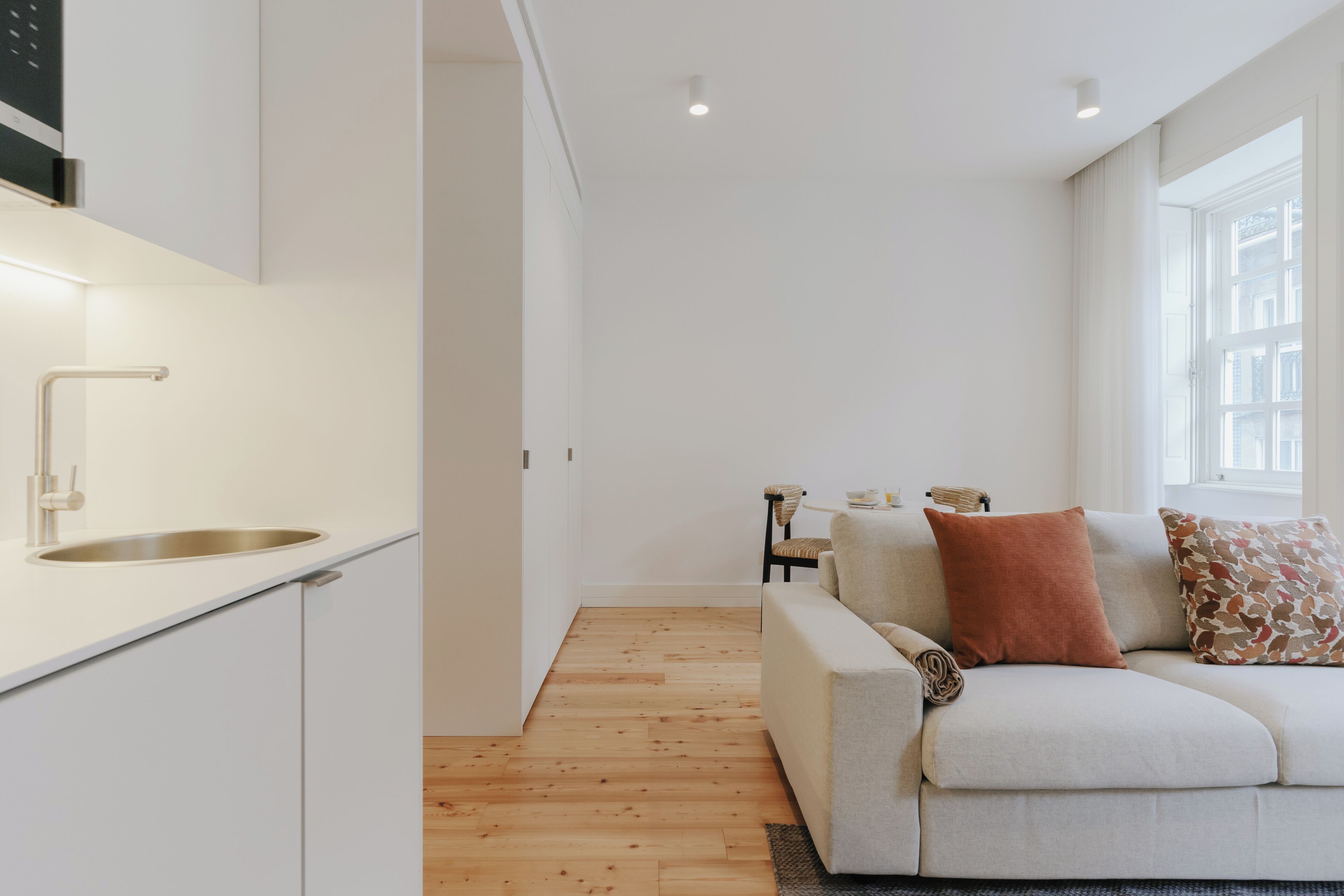Eine moderne Küche mit Spüle aus Edelstahl und Holzboden. Im Wohnbereich befindet sich ein weißes Sofa mit einem braunen Kissen und einem gemusterten Kissen.