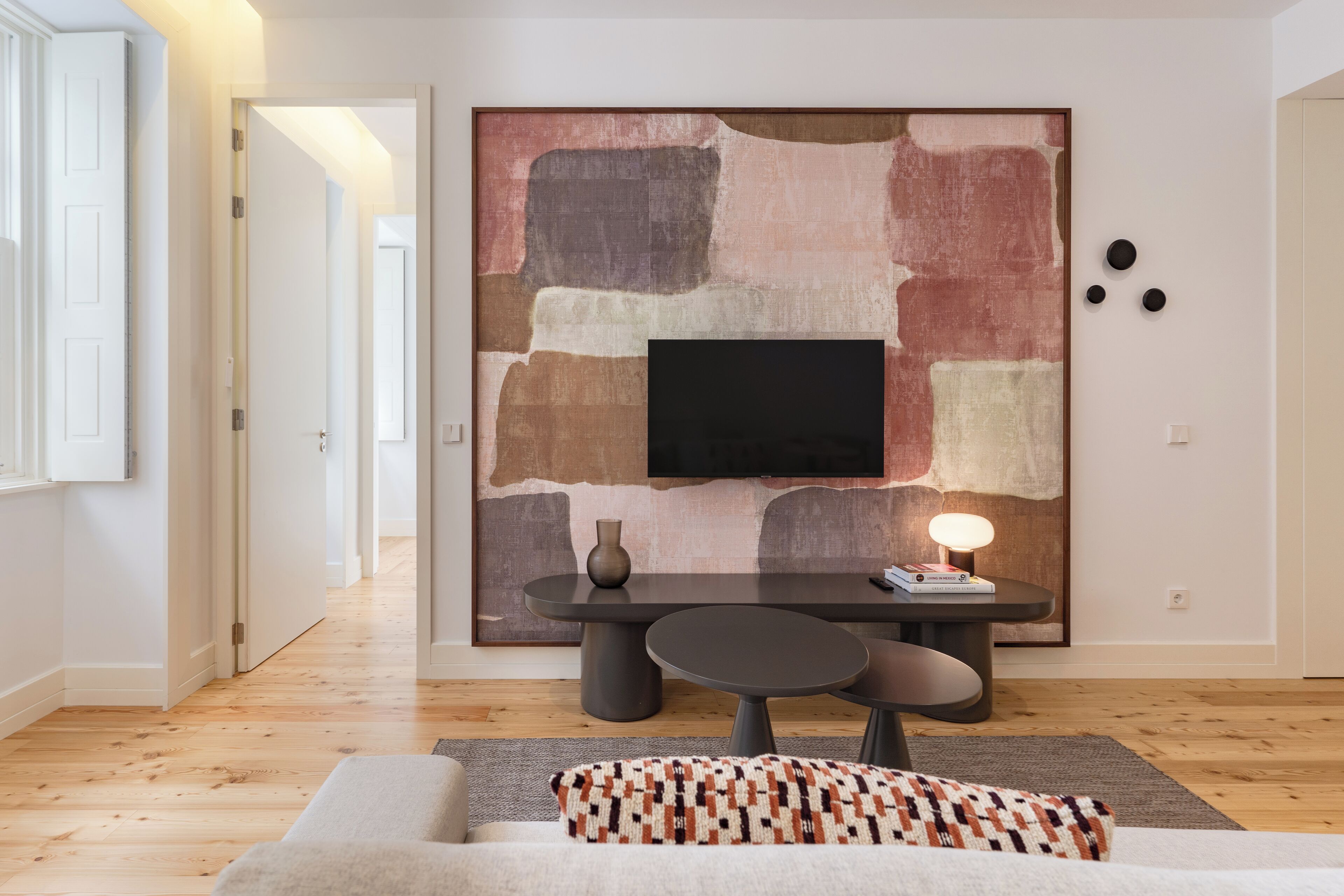 A modern living room with a flat-screen TV mounted on a textured wall, a dark wooden dining table with chairs, and a patterned throw pillow on a sofa.
