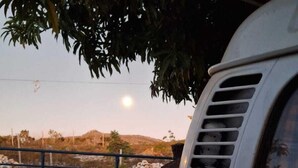 A red vehicle parked outdoors with a metal fence and a tree in the foreground. - Casa Donjá (Serro)