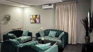 A living room with two green sofas, a small table, a painting, an air conditioning unit, and a curtain. - Dankani Guest Palace Hotel (Sokoto)