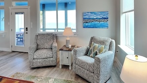 A living room with two patterned armchairs, a small table with a lamp, a painting on the wall, and a view of the outdoors through a large window. - Heron House | Pet-friendly with Private Pool (Rockport)