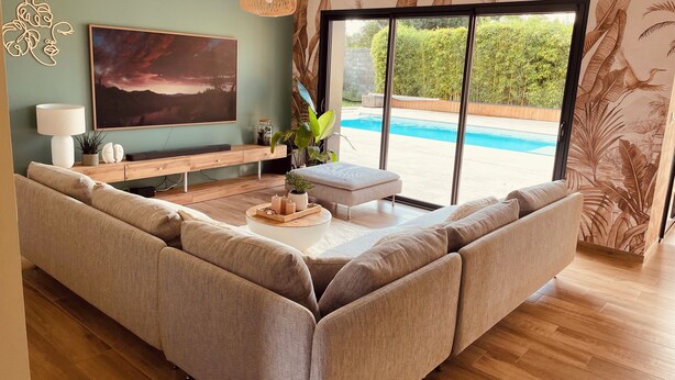 En moderne stue med en stor sofa, et sofabord og et fjernsyn. Der er et vindue med udsigt til en swimmingpool og grønne omgivelser.