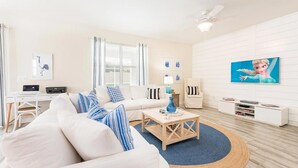 A modern living room with a white sofa, blue and white pillows, a wooden coffee table, and a TV on the wall. - South-Facing Pool & Spa • Game Room • Conservation View • Near Disney (Four Corners)