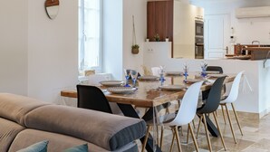 Ein modernes Wohnzimmer mit einem Holzespeisetisch, gedeckt für sechs Personen, umgeben von weißen und schwarzen Stühlen. Ein beiges Sofa mit blauen Kissen befindet sich im Vordergrund.
