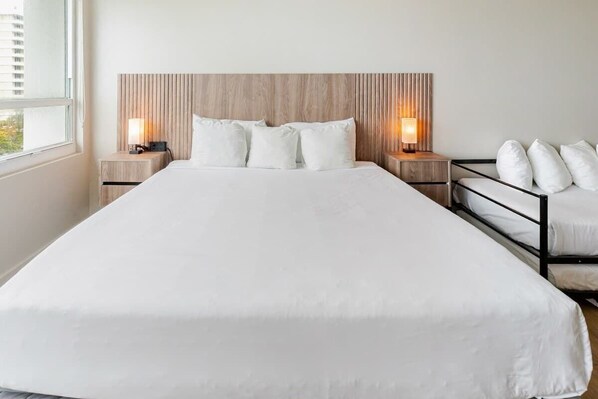 A large bed with a wooden headboard, two bedside tables with lamps, and a view of a cityscape through a window. - Shine Suites Studio (Miami)