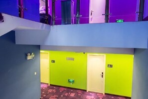Un couloir d’hôtel avec un plafond bleu, des murs verts et une moquette rouge. On y trouve des portes avec des panneaux indiquant les numéros de chambre.