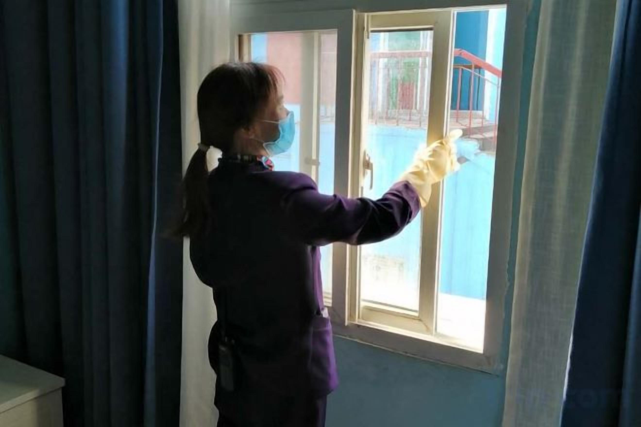 A person standing by a window, wearing a purple outfit and a face mask, holding a yellow object.