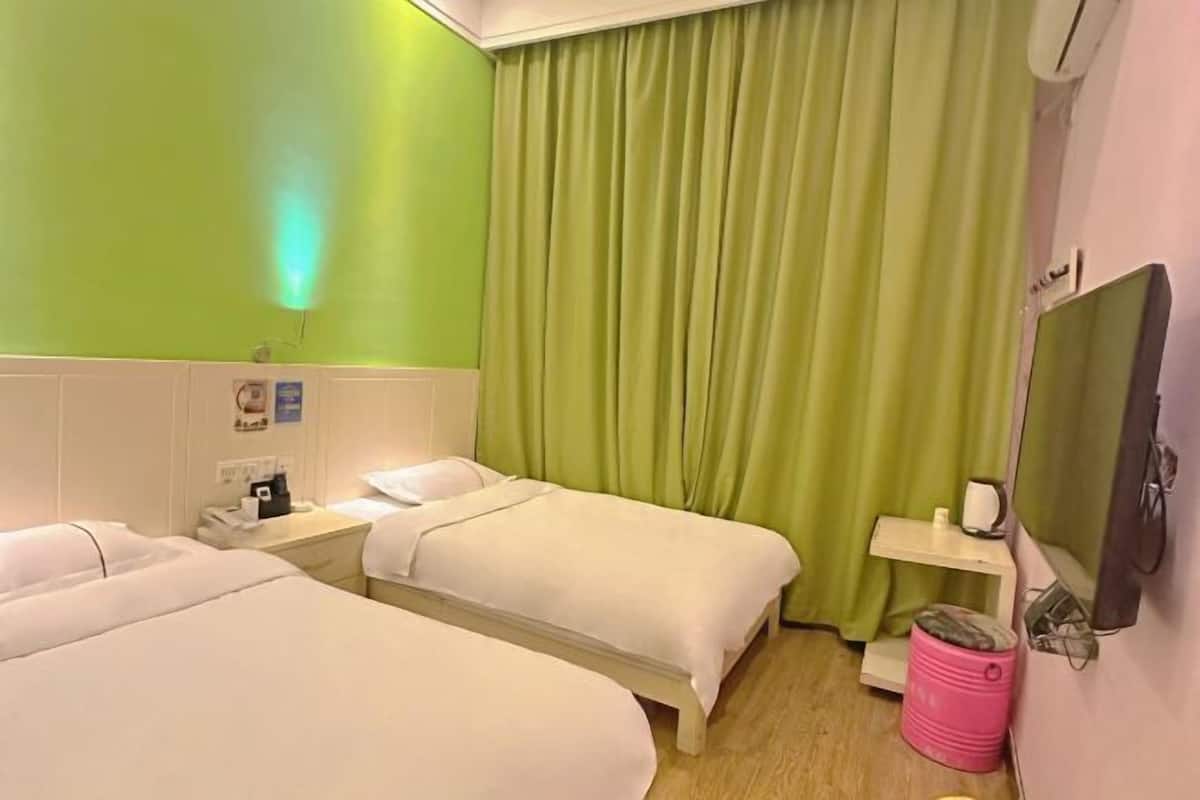 Habitación de hotel con dos camas, una mesita de noche, un televisor, una cortina verde y un cubo de basura rosa.