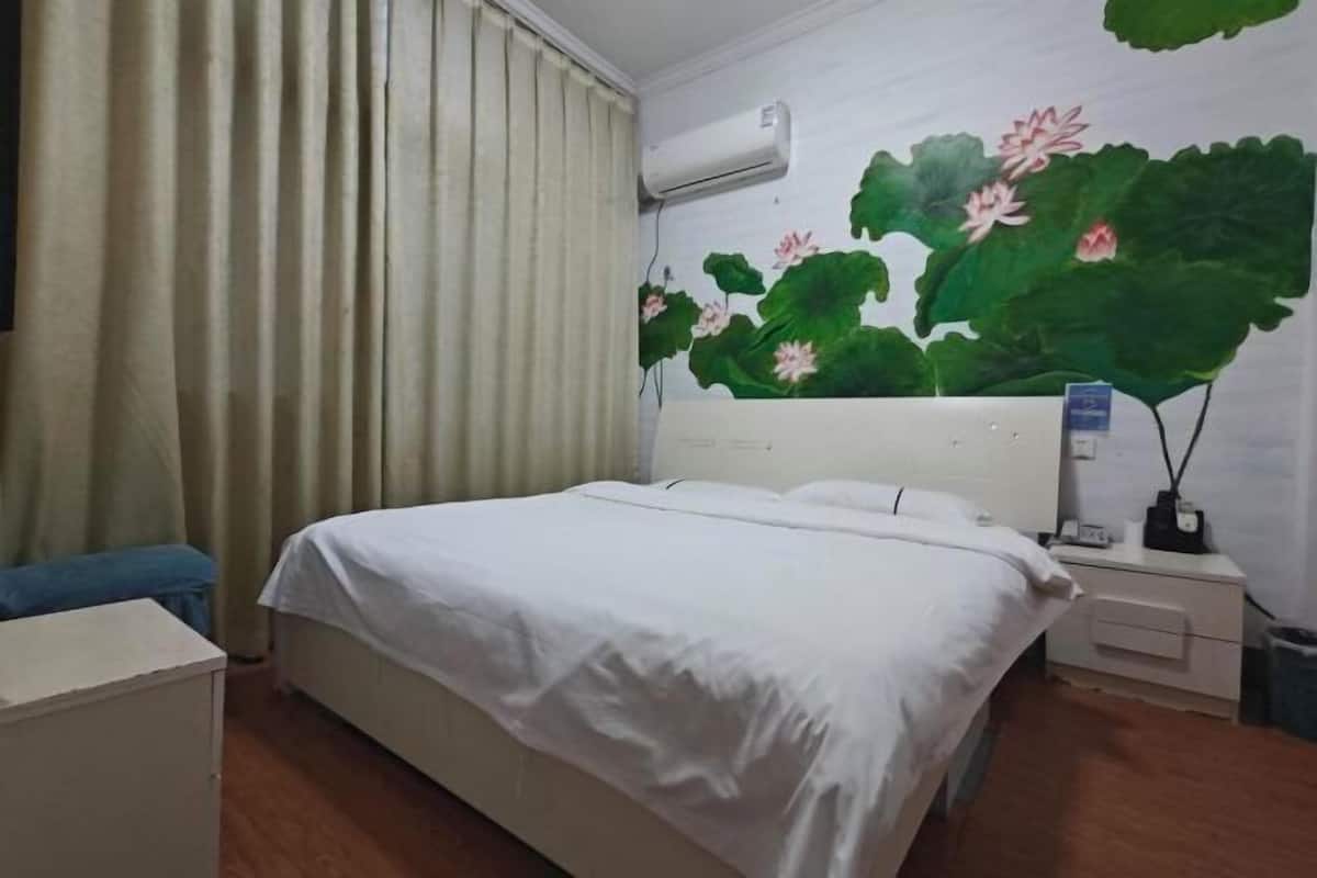 Una habitación de hotel con cama, cortinas, unidad de aire acondicionado, mesita de noche y un mural de flor de loto en la pared.