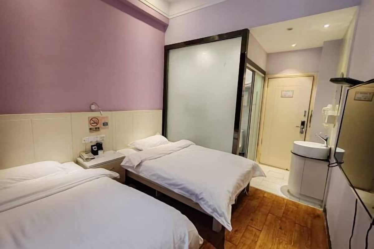 Habitación de hotel con dos camas, una mesita de noche con teléfono, una lámpara de pared y una puerta que da a un baño con lavabo e inodoro.