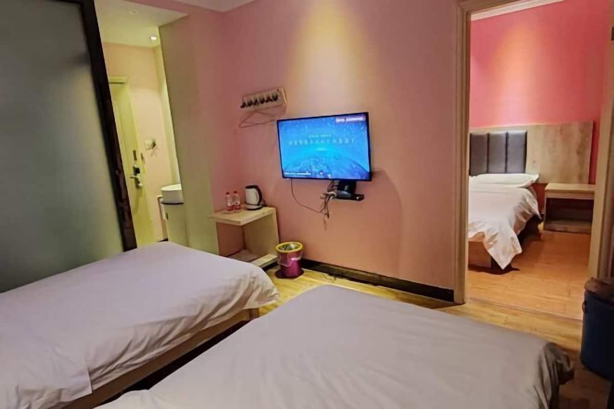 Habitación de hotel con dos camas, un televisor montado en la pared, una mesita con una lámpara y un cubo de basura.