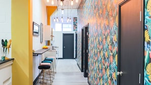 A narrow hallway with a kitchen area on the left, featuring bar stools and a counter. The walls are decorated with a tropical leaf pattern, and there is a door at the end of the hallway.