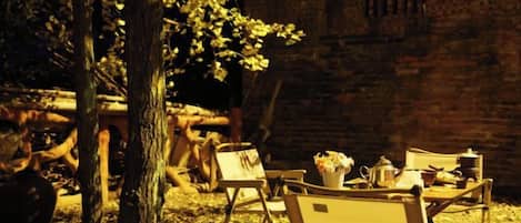 An outdoor seating area with a table set for two, surrounded by chairs, under a tree.