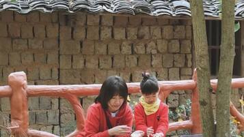 Two individuals seated on a wooden bench outdoors, with a brick wall and tiled roof in the background.
