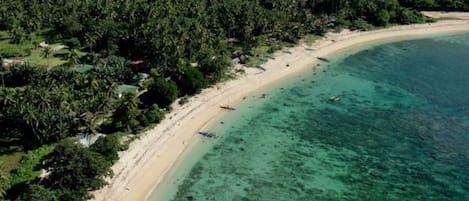 Vista aérea de una playa con aguas turquesas cristalinas, rodeada de exuberante vegetación tropical y algunas construcciones.