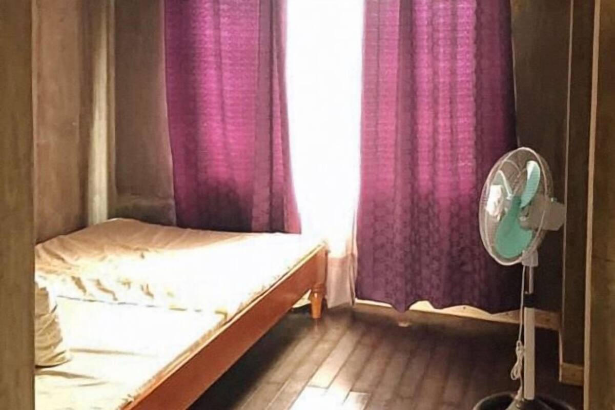 A room with a bed, a standing fan, and curtains.