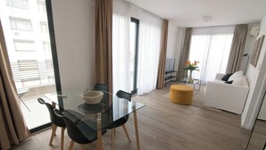 Une chambre d’hôtel moderne avec une table à manger en verre, des chaises noires, un pouf jaune, un canapé blanc et une grande fenêtre avec des rideaux.