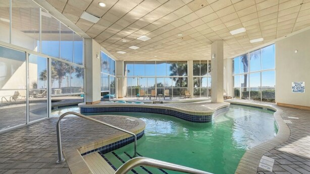 An indoor swimming pool with clear glass walls, a tiled floor, and a pool area with lounge chairs. - Carolinian 1732-Oceanfront 2BR/2BA-Pool (Myrtle Beach)
