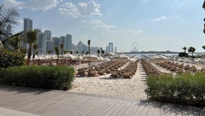 En strandpromenad med rader av solstolar och parasoller, palmer och en stadsbild i bakgrunden.
