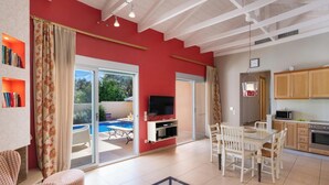 Un vaste espace de vie agrémenté d’un mur rouge, une porte-fenêtre coulissante donnant sur une piscine, un coin repas avec une table et des chaises, et une cuisine dotée de meubles en bois.