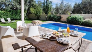 Un espace repas aménagé au bord de la piscine, avec une table dressée pour le petit-déjeuner, des chaises et une vue sur la piscine et la végétation environnante.