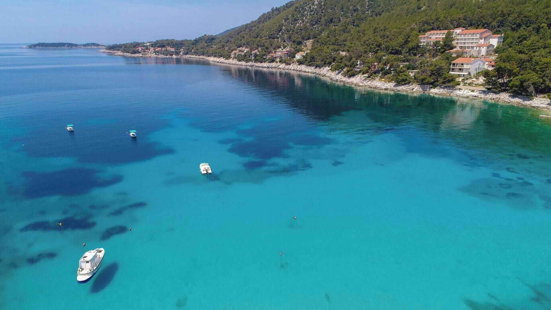 Une zone côtière aux eaux turquoise et limpides, où l’on aperçoit plusieurs bateaux et un ensemble de bâtiments situés sur le flanc d’une colline.