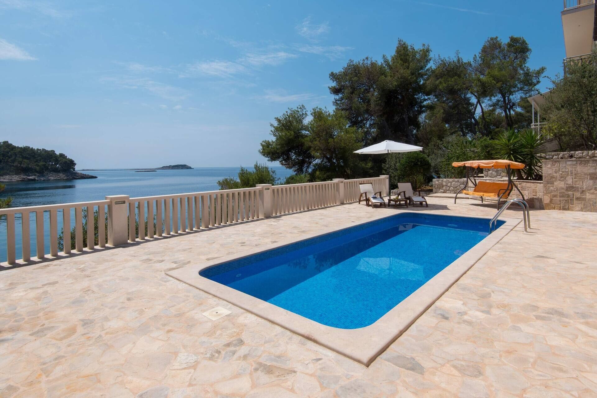 A swimming pool with a stone deck, surrounded by lounge chairs and umbrellas, overlooking a coastal area with trees and a clear sky.