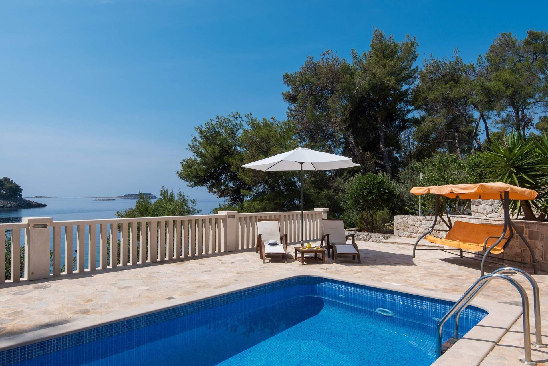 Área da piscina com vista para o mar, um guarda-sol branco e cadeiras de descanso.