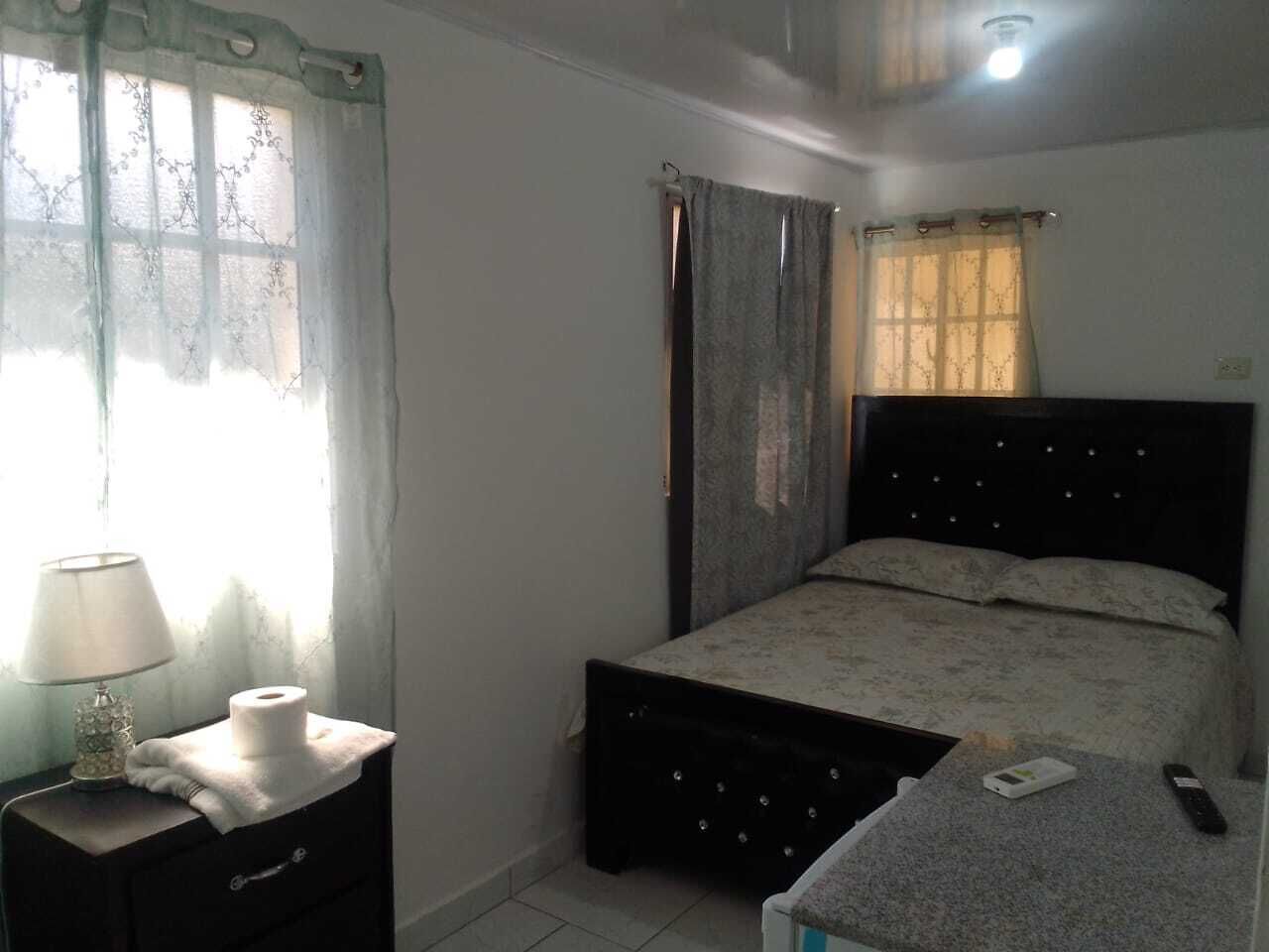 Un dormitorio con cama, mesita de noche con lámpara, control remoto y ventana con cortinas.