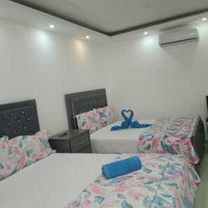 A hotel room with two beds, each with floral-patterned bedding and a heart-shaped towel arrangement. There are bedside tables with remote controls, and an air conditioning unit mounted on the wall. - Family city hotel (Puerto Plata)