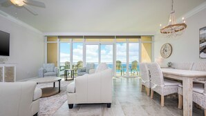A spacious living room with a white sofa, a coffee table, and a dining area with wicker chairs. Large windows offer a view of the outdoors.