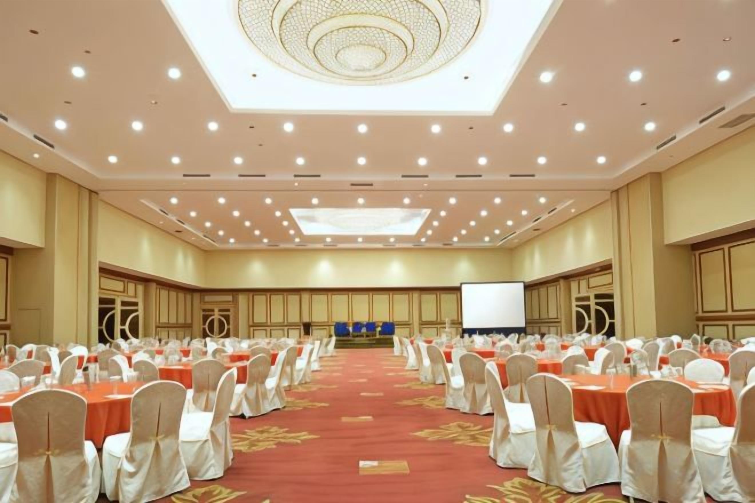 A large conference room with rows of tables and chairs, a stage with a screen, and a ceiling with recessed lighting.