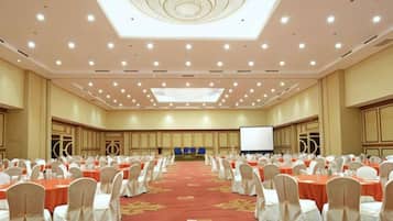 A large conference room with rows of tables and chairs, a stage with a screen, and a ceiling with recessed lighting.