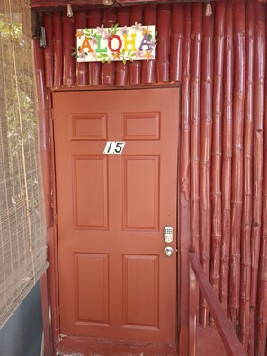 Une porte marron portant le numéro « 15 » et dotée d’un clavier pour l’ouverture, le tout adossé à un mur en bambou sur lequel est affiché un panneau indiquant « ALOHA ».